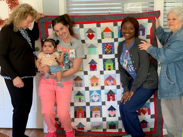 Four women are standing in front of a quilt featuring little houses. One of the women holds a small child.